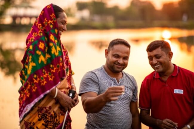 Dos hombres están de pie en un lago sonriendo junto a una mujer. Todos miran algo que sostiene el hombre del medio, al atardecer.