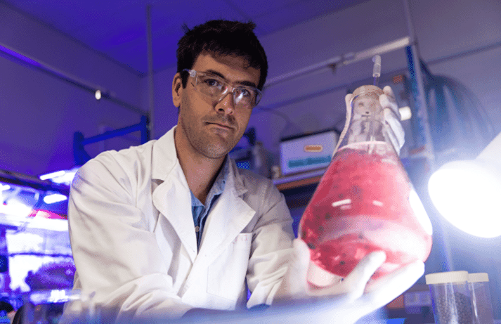 a man in a white coat holding a beaker of seaweed in a laboratory