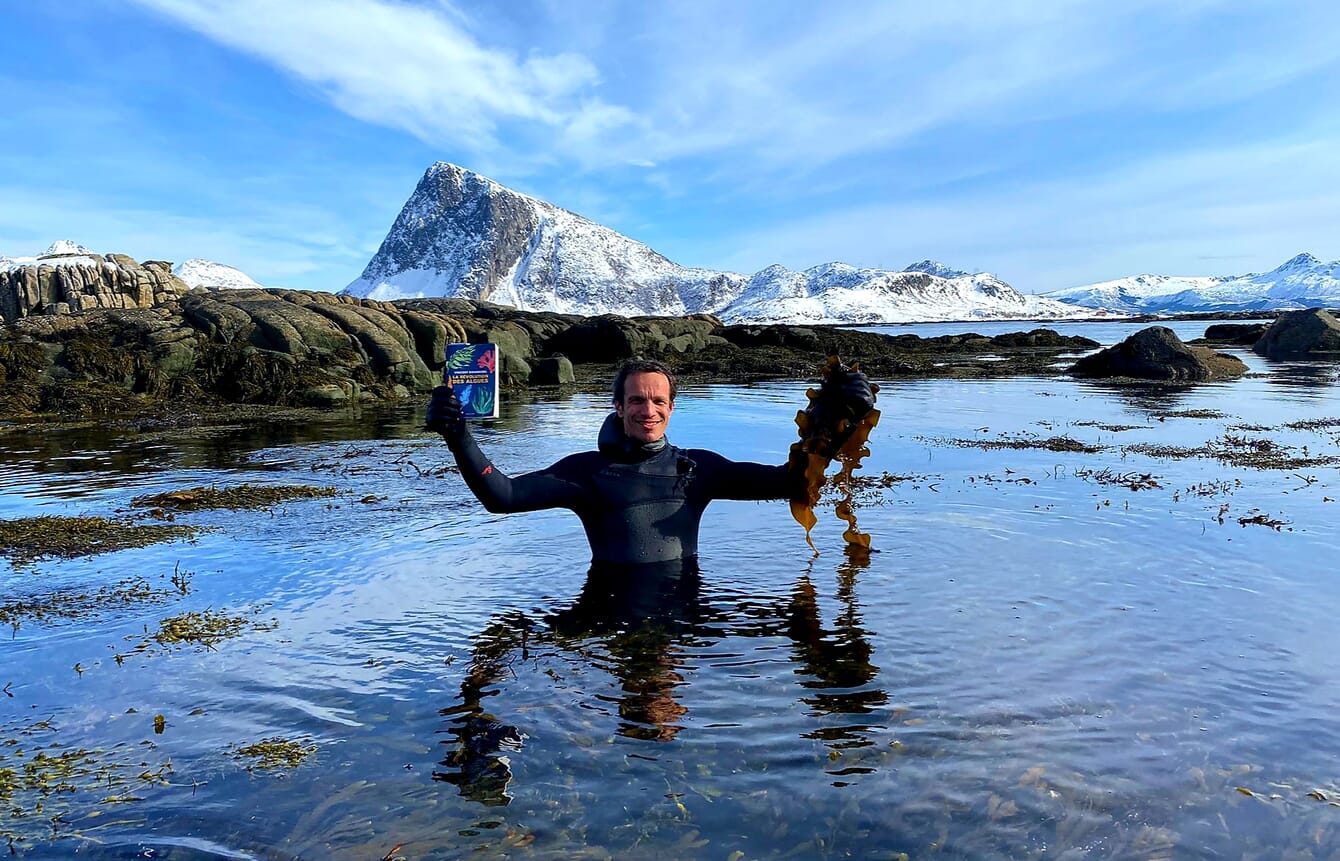 Man standing in water holding a book in one hand and seaweed in the other hand