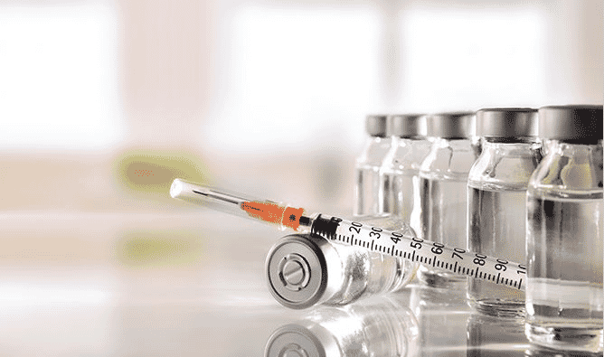 A loaded syringe with vials of vaccines in the background