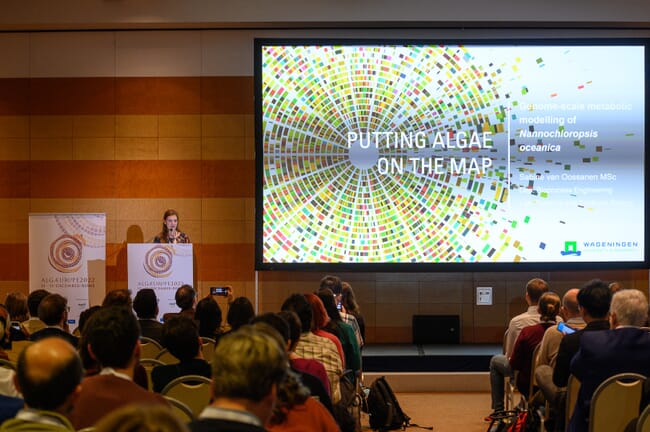 A conference room full of seated guests, a woman standing to left at a podium speaking and a projector screen with a slide saying, putting algae on the map.