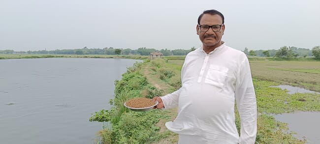 A man with a plate of feed standing beside a pond.