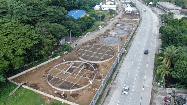 um canteiro de obras é retratado próximo a uma estrada, enquanto um incubatório de camarões é mostrado no meio da construção.