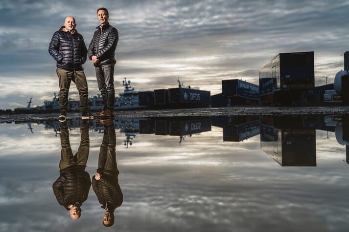 two men, reflected in a puddle, standing in front of an industrial backdrop