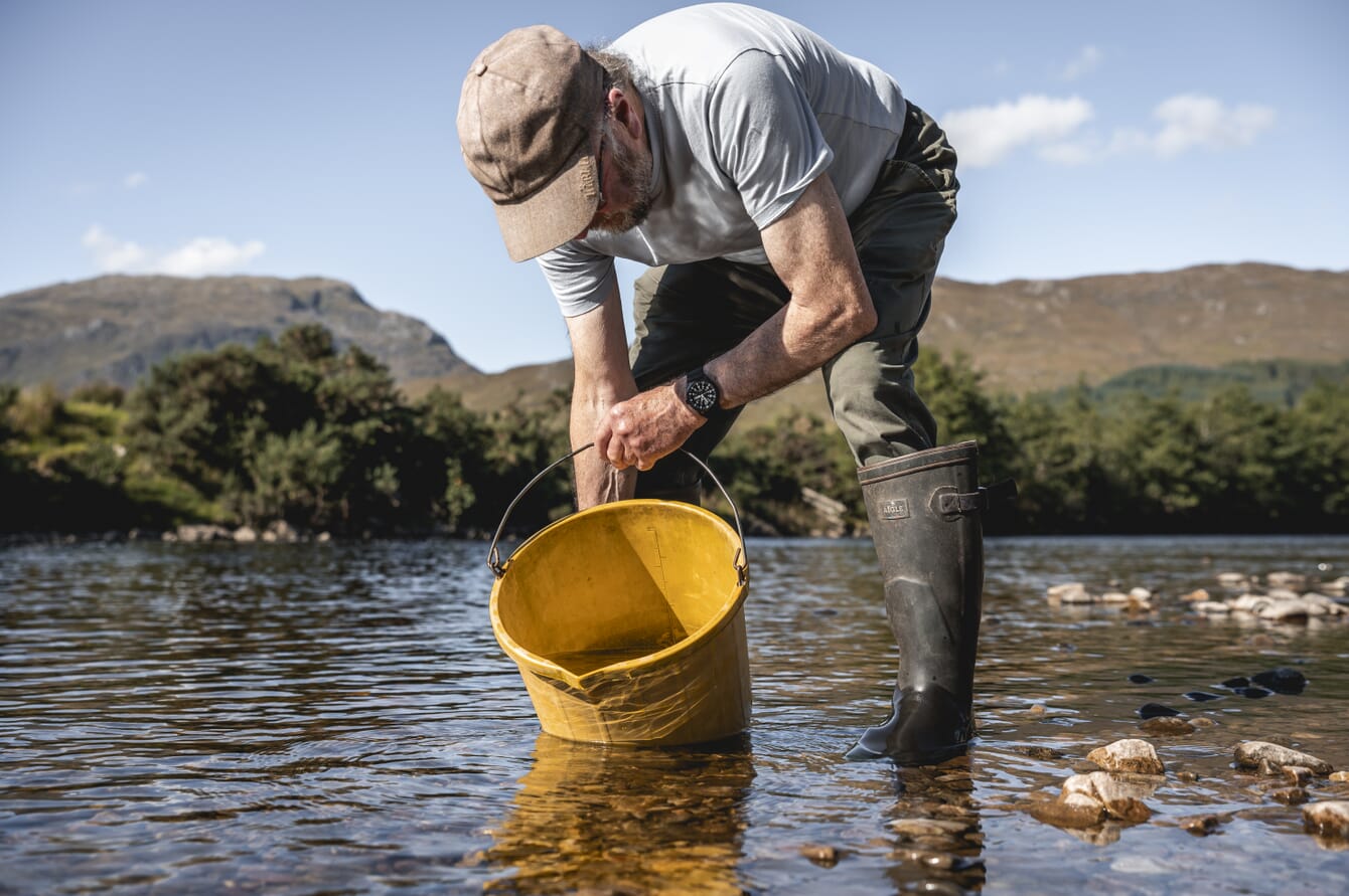 A scientist releasing salmon fry into a river in Scotland on a sunny day.