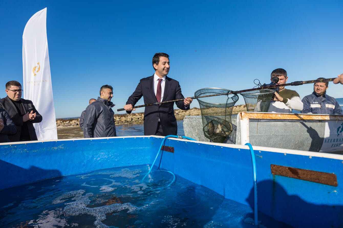 Um homem de terno segurando um peixe em uma rede enquanto é filmado.