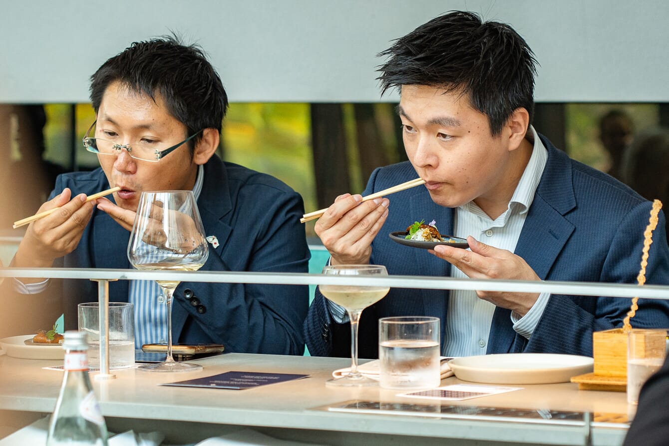 Two men in suits eating with chopsticks.