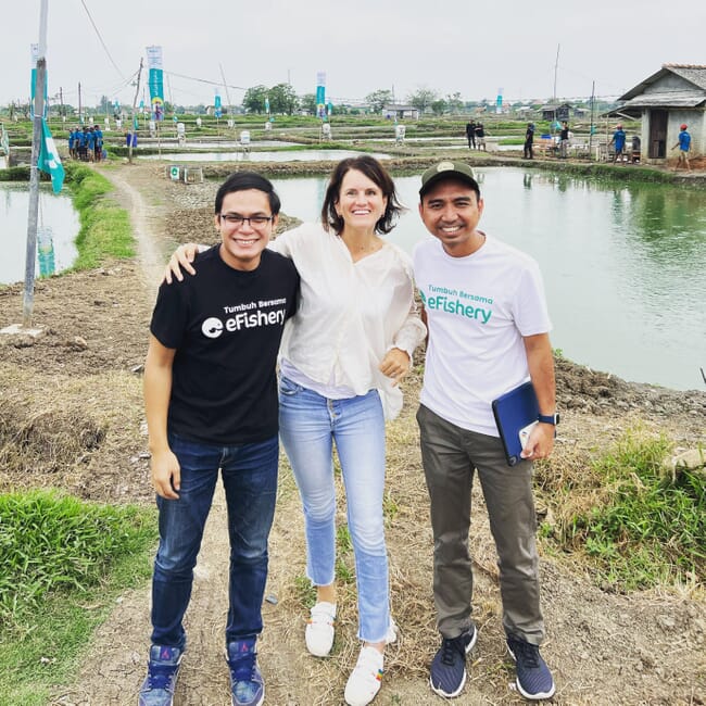 Tres personas sonriendo a la cámara delante de estanques de camarones